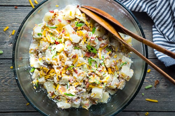 Loaded potato salad in large glass mixing bowl with wooden salad tongs on gray wood surface
