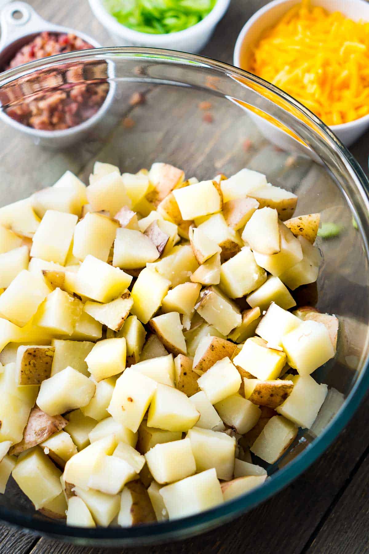 cooked diced potatoes in glass bowl next to potato salad ingredients in small bowls