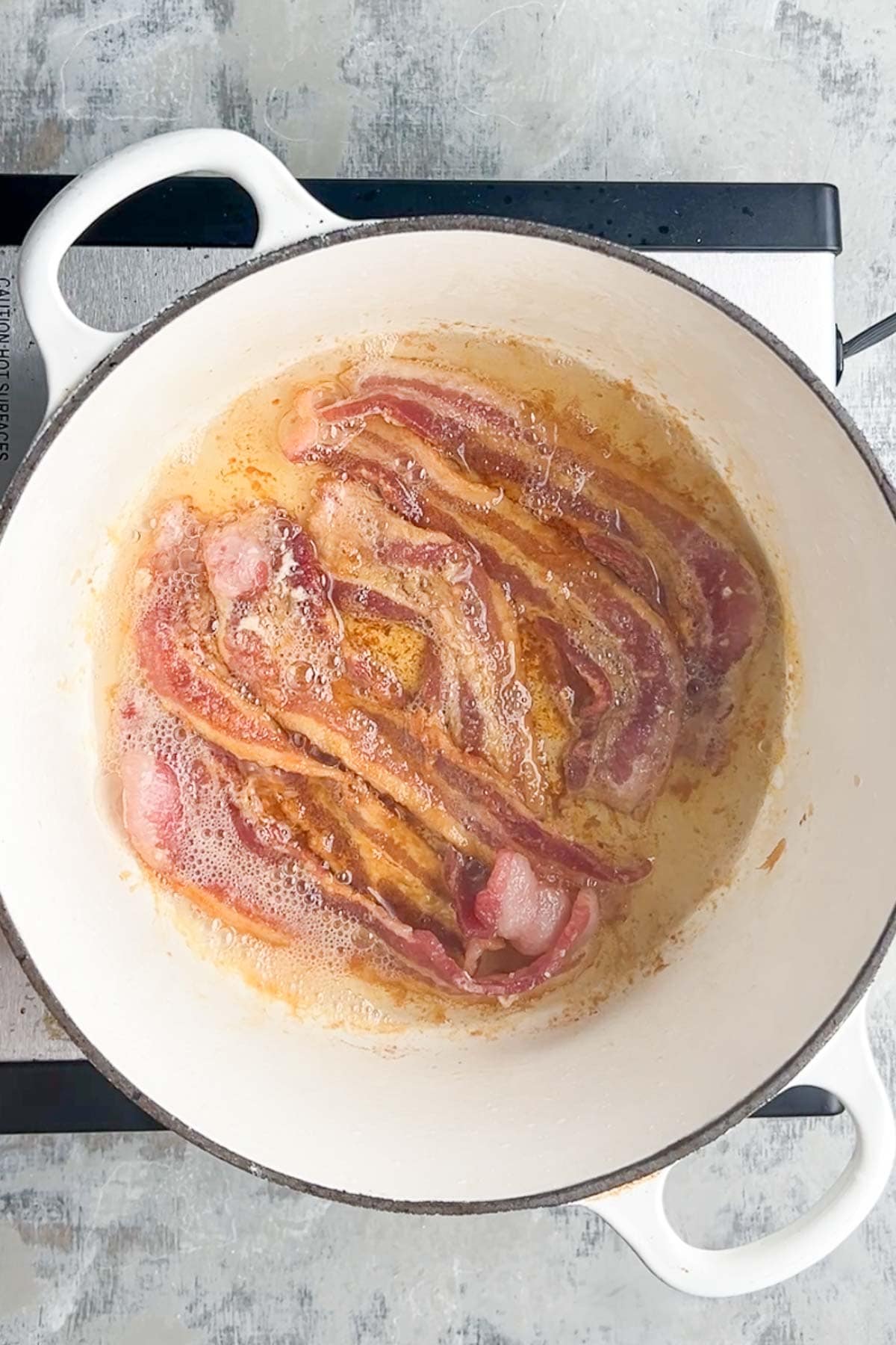 Strips of bacon frying in a white cast iron pot on a stovetop, with grease and foam visible around the bacon&mdash;perfect for starting a rich loaded baked potato soup.