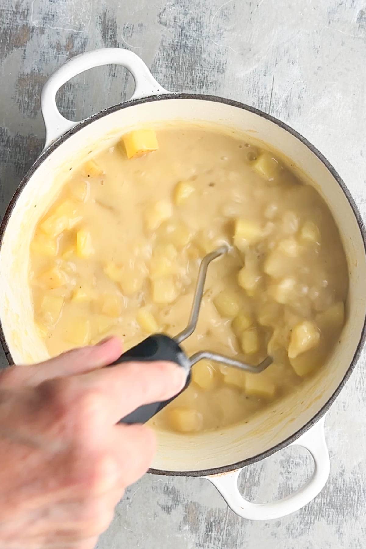 A hand uses a potato masher to mash cooked potatoes in a white pot, preparing the base for a hearty loaded baked potato soup.