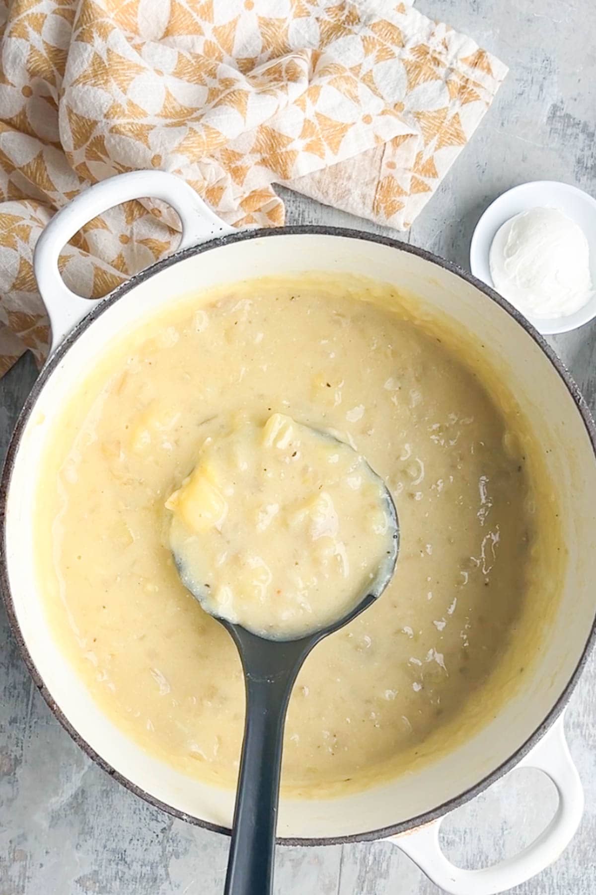 A pot of creamy loaded baked potato soup with a ladle, set beside a patterned cloth napkin and a small dish of sour cream.