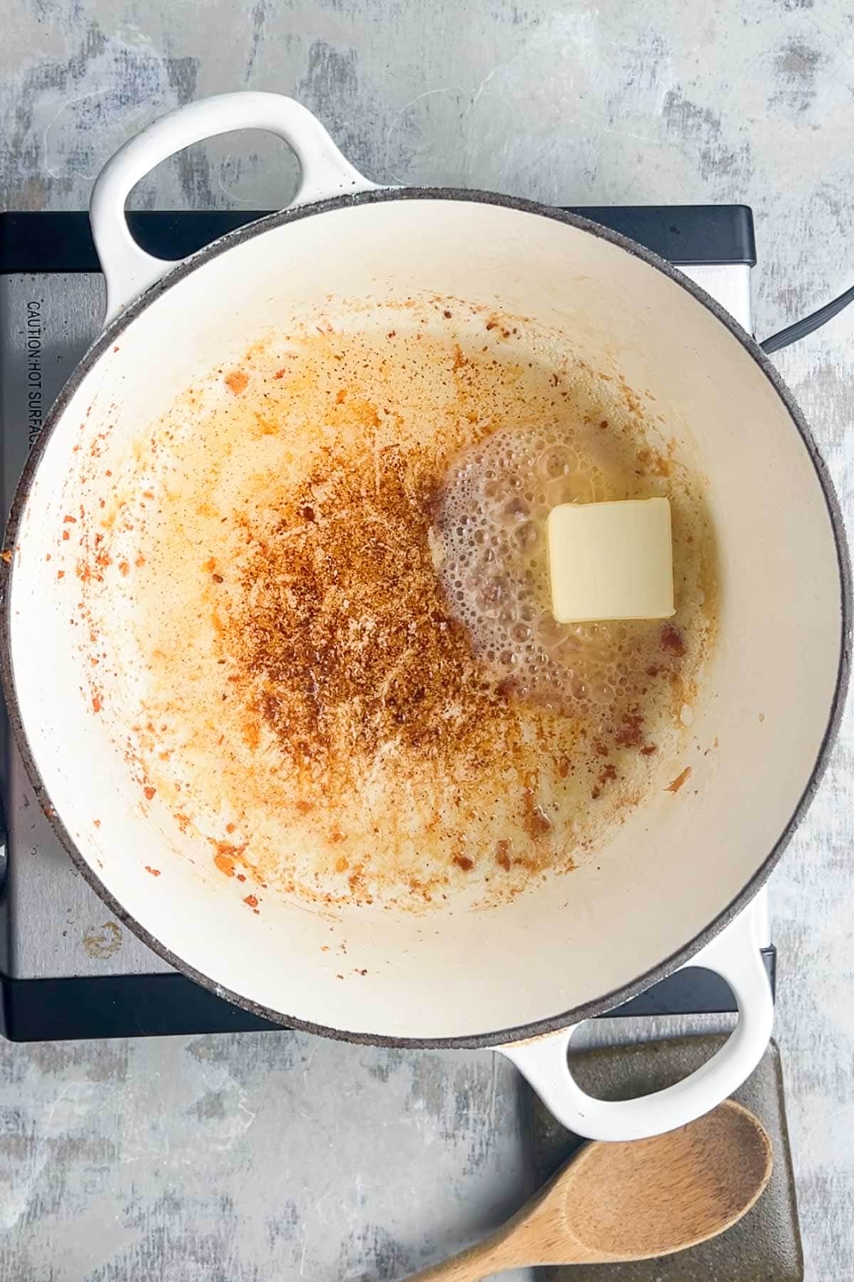 A white Dutch oven on a stovetop contains melted brown bits and a melting pat of butter, setting the stage for delicious loaded baked potato soup; a wooden spoon rests nearby.