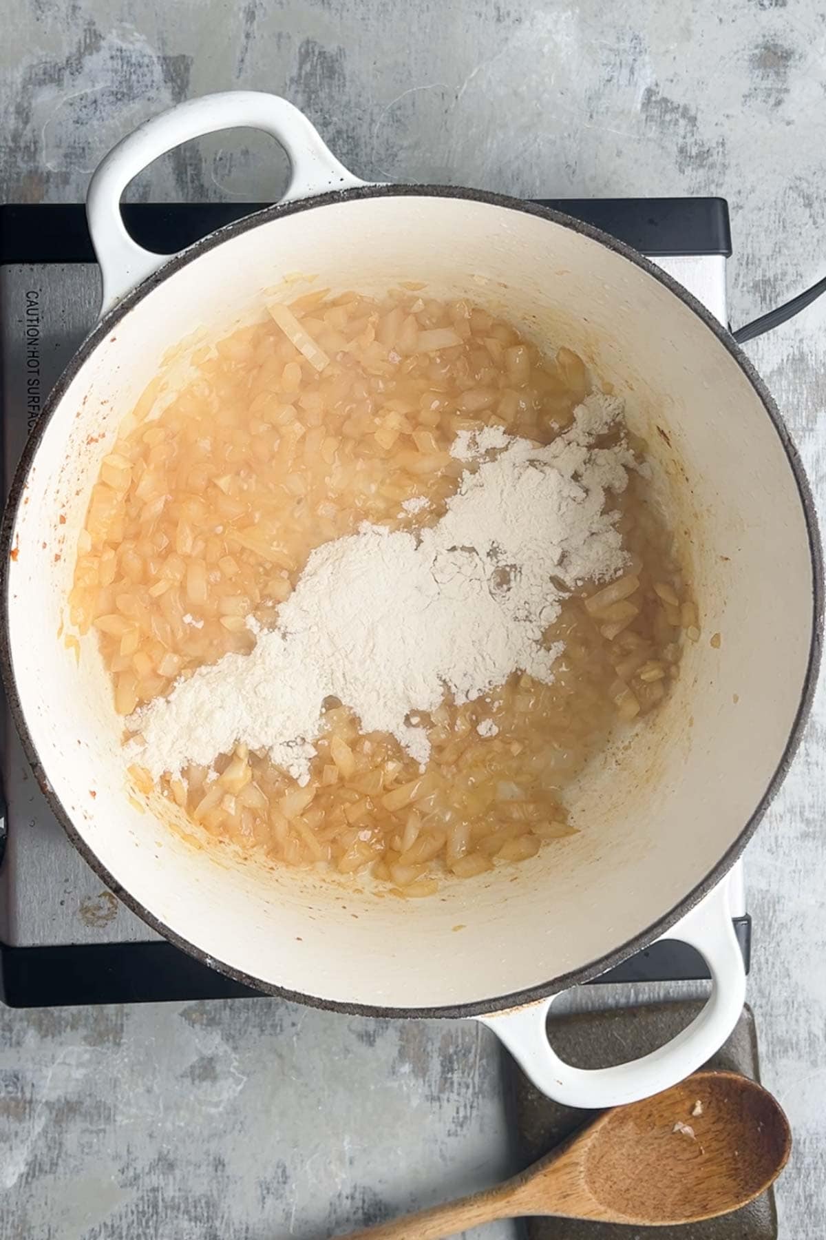 Chopped onions cooking in a white pot on the stovetop, with a heap of flour added&mdash;steps away from becoming creamy loaded baked potato soup; a wooden spoon rests nearby.