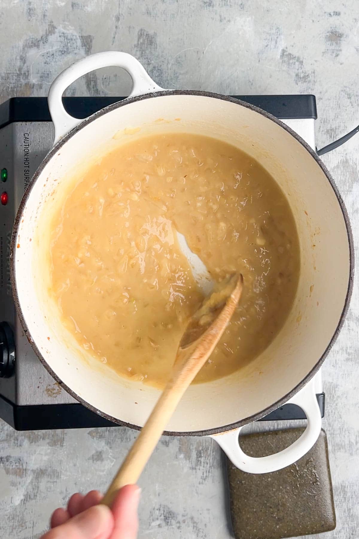 A hand stirs creamy onions and broth in a white pot on a stovetop with a wooden spoon to make loaded baked potato soup.