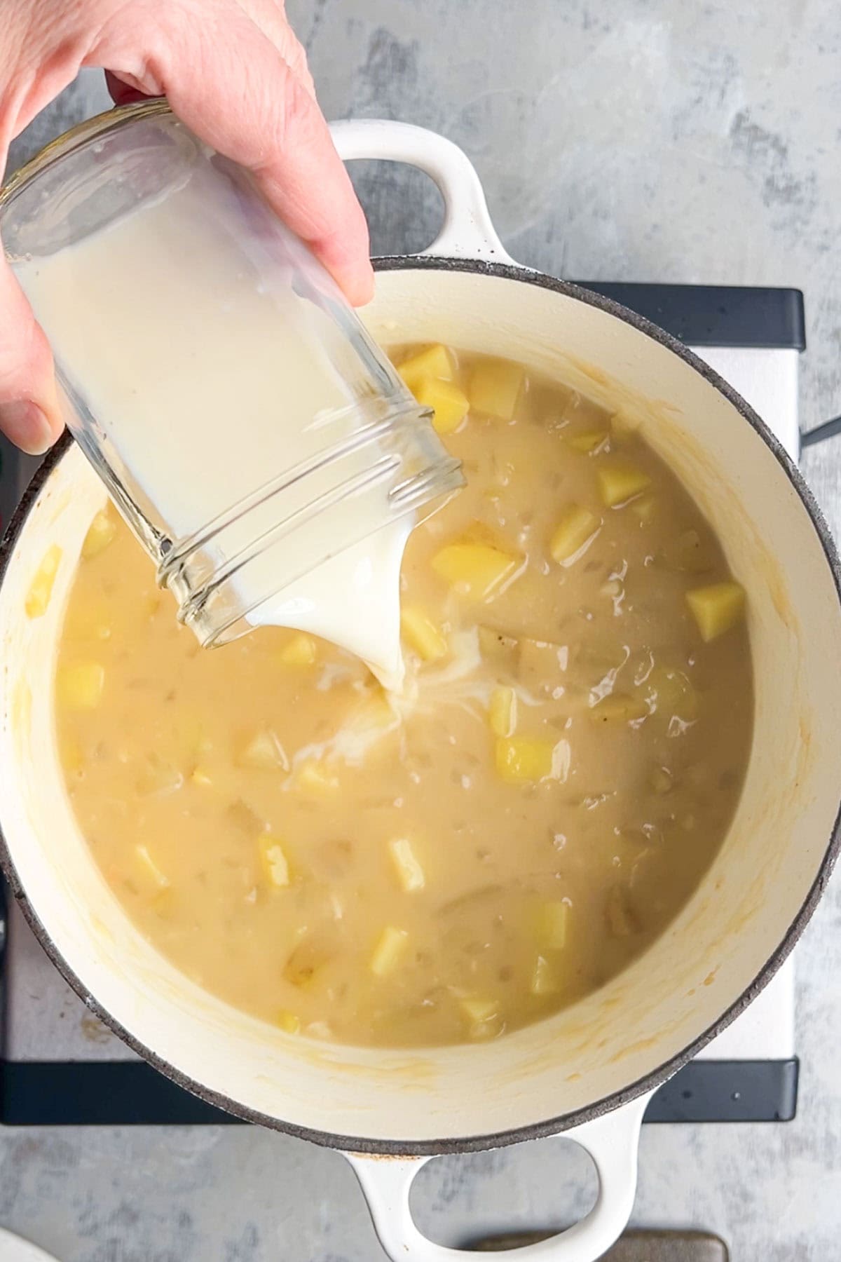 A hand pours a milk from a glass into a pot of loaded baked potato soup with diced potatoes simmering on the stovetop.