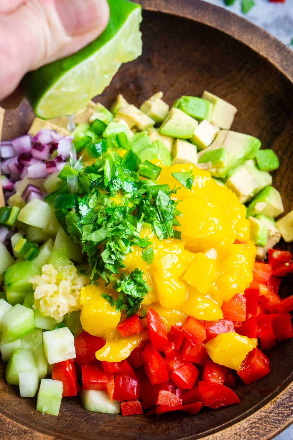 hand squeezes fresh lime over ingredients for avocado mango salsa in brown wood bowl