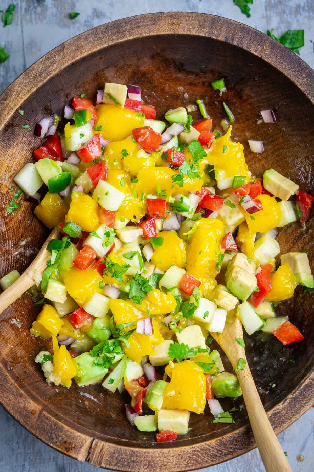 ingredients for avocado mango salsa have been tossed in brown wood bowl with two wooden spoons