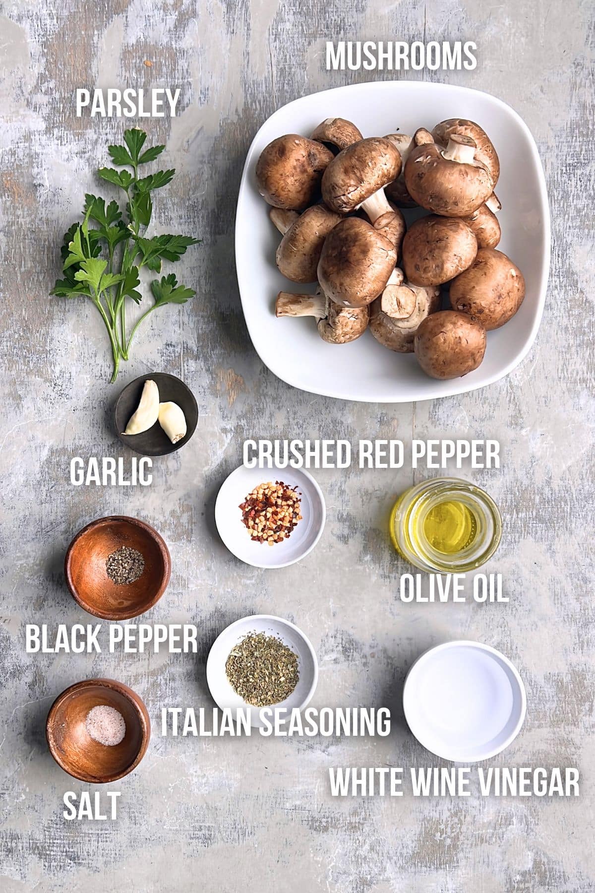 Flat lay of ingredients for marinated mushrooms, including parsley, mushrooms, garlic, crushed red pepper, olive oil, black pepper, Italian seasoning, salt, and white wine vinegar.
