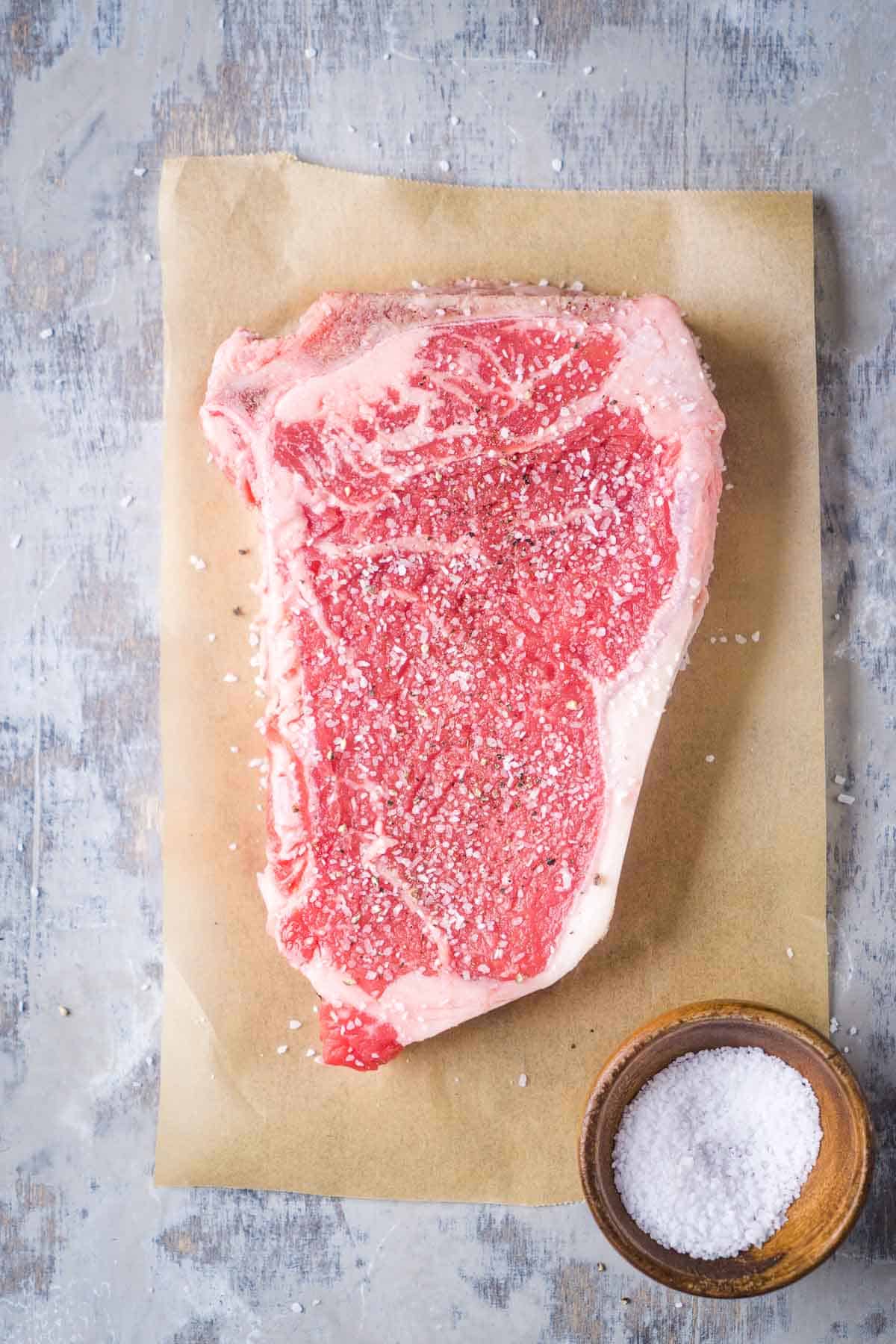 uncooked strip steak on butcher paper next to pinch bowl of salt