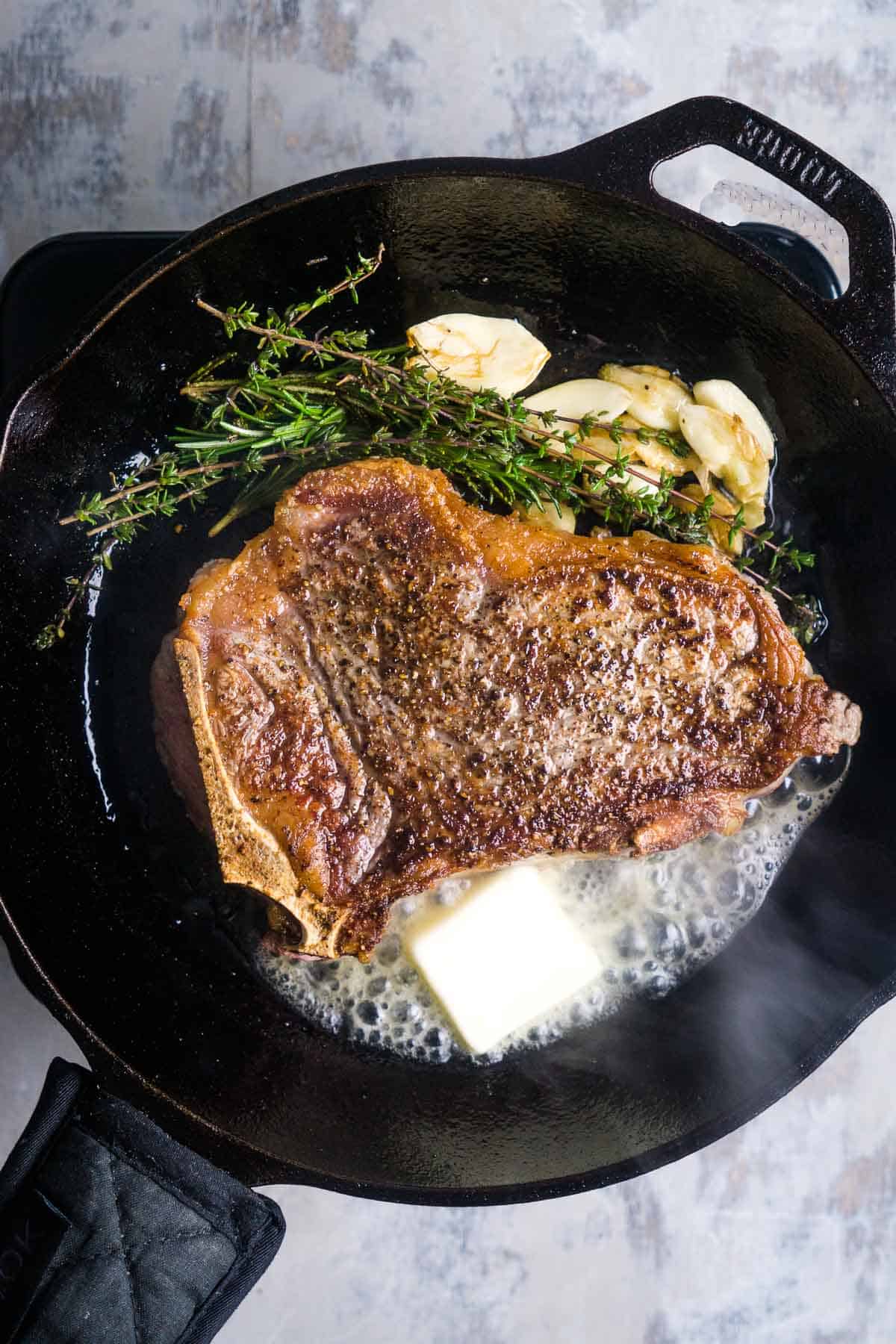 juicy, seared New York strip steak in cast iron skillet with garlic cloves, herb sprigs, and pat of melting butter
