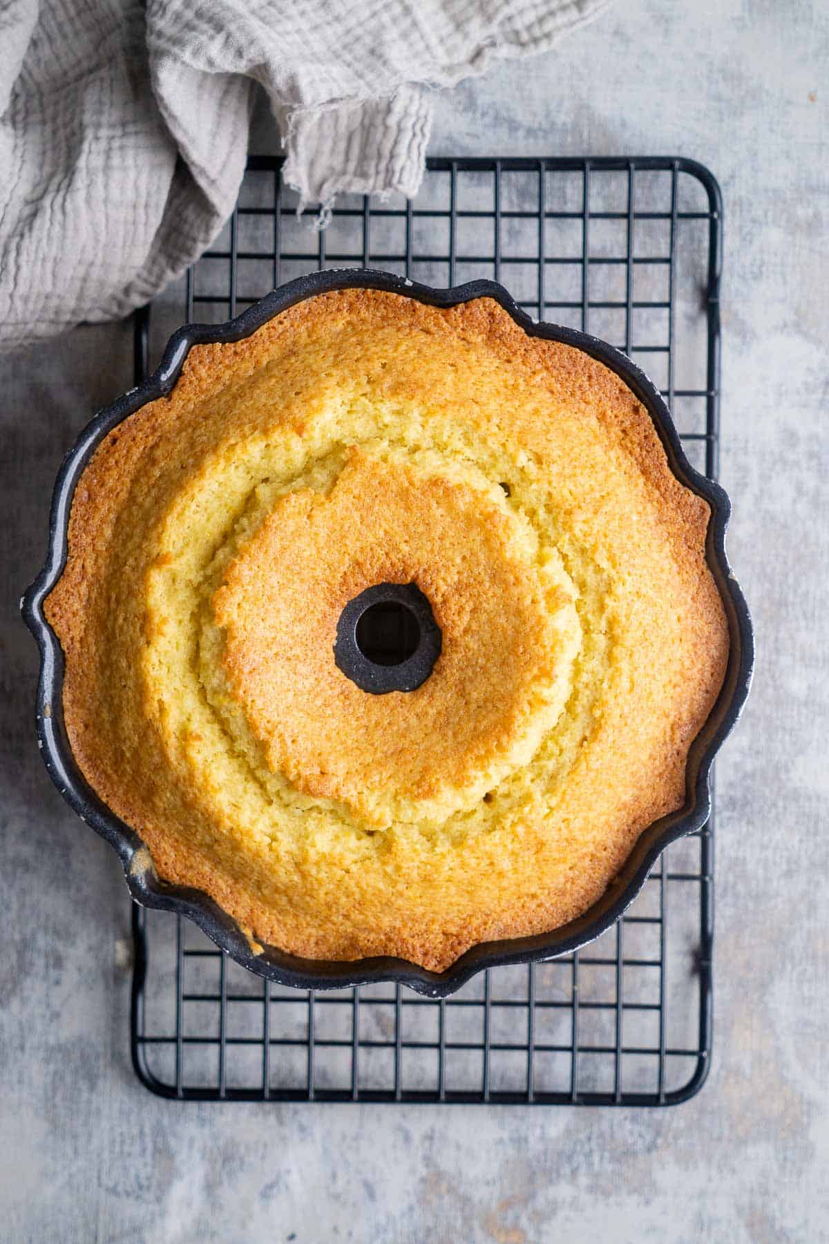 Golden brown orange olive oil cake in a bundt cake pan on a cooling rack next to gray linen.