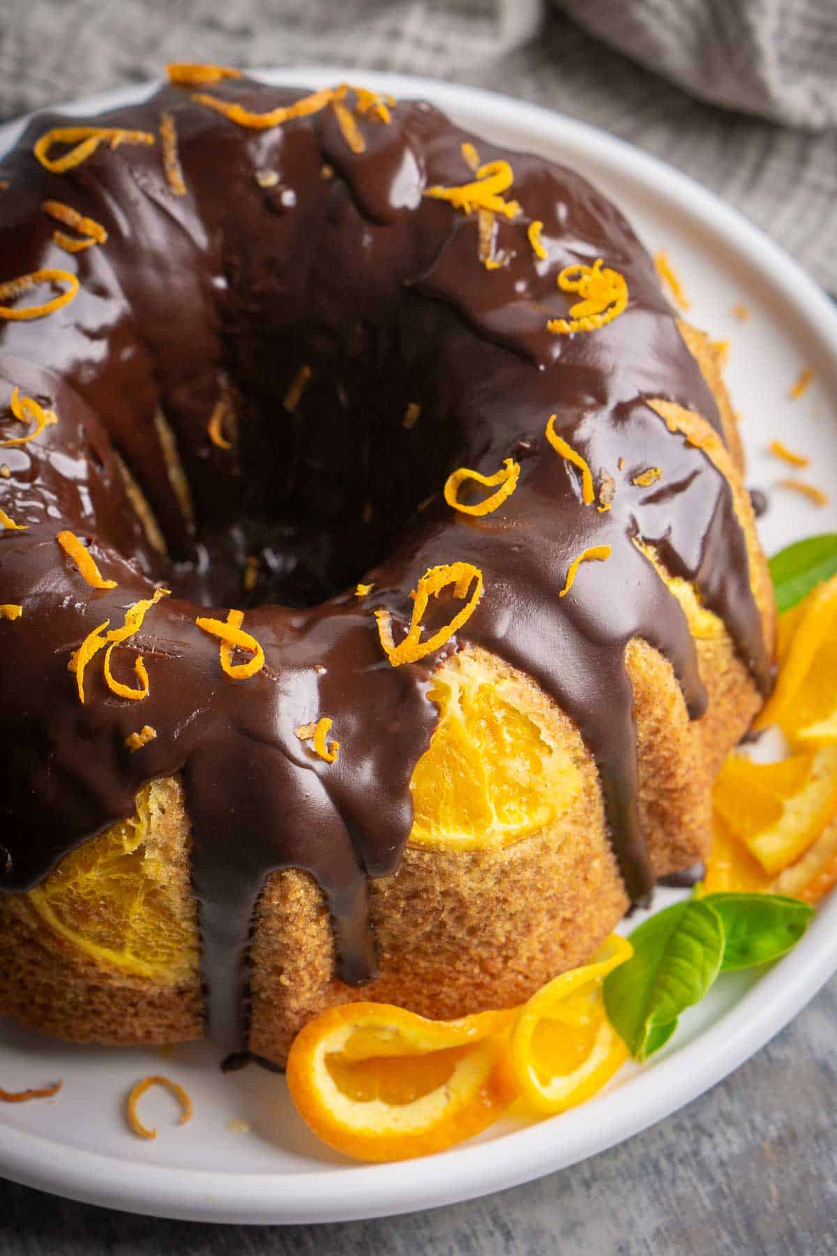 Orange olive oil cake on white plate with chocolate ganache topping and orange zest, garnished with folded fresh orange slices.