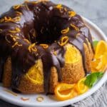 Orange olive oil cake on white plate with chocolate ganache topping and orange zest, garnished with folded fresh orange slices.