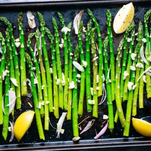 baked asparagus on dark colored baking sheet with garlic, lemon wedges, and shallots.