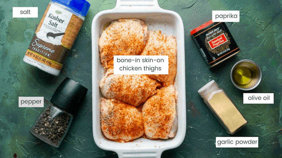 seasoned uncooked chicken thighs in white baking dish next to containers of salt, pepper, paprika, oil, and garlic on green background