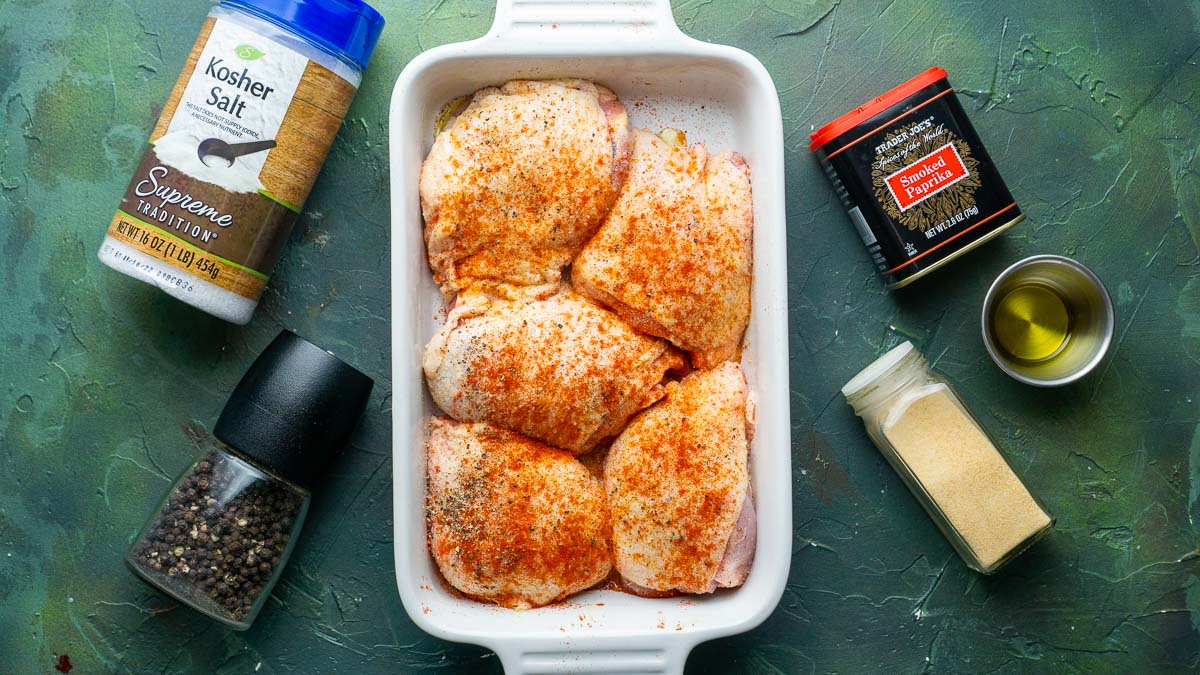 Five raw chicken thighs in a white dish, seasoned with paprika, surrounded by containers of kosher salt, black pepper, garlic powder, paprika, and a small bowl of olive oil on a green surface.
