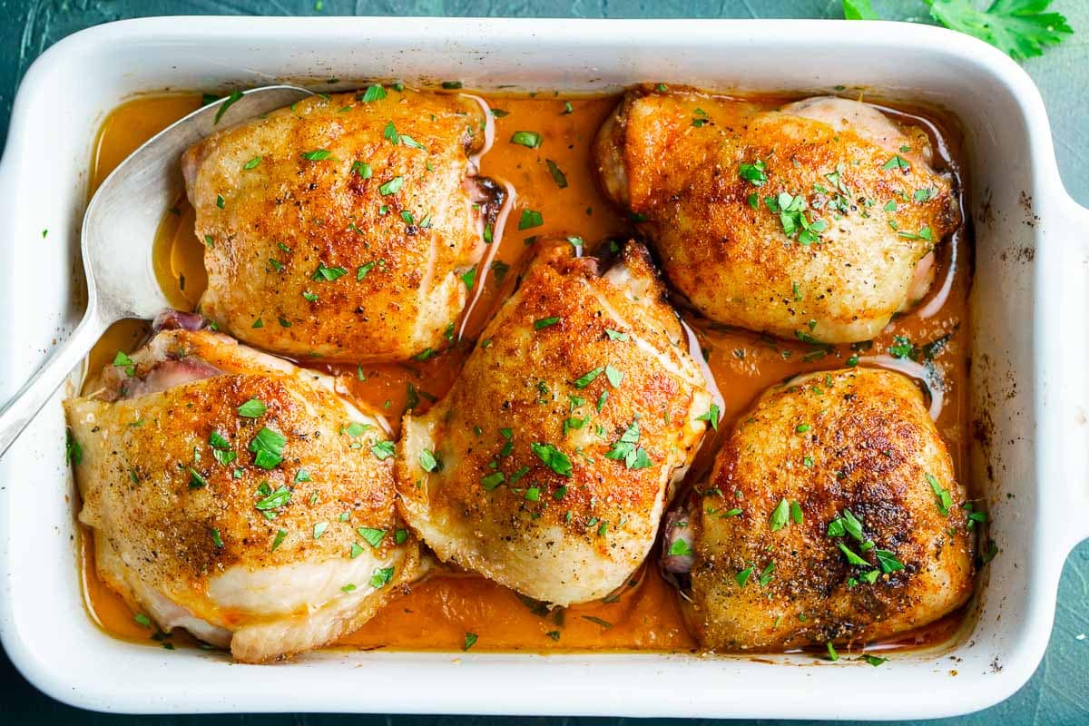 Five baked chicken thighs with golden, crispy skin in a white baking dish with juices and a serving spoon, garnished with chopped parsley.