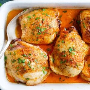 Baked chicken thighs with golden, crispy skin in a white dish, garnished with chopped parsley and surrounded by cooking juices.