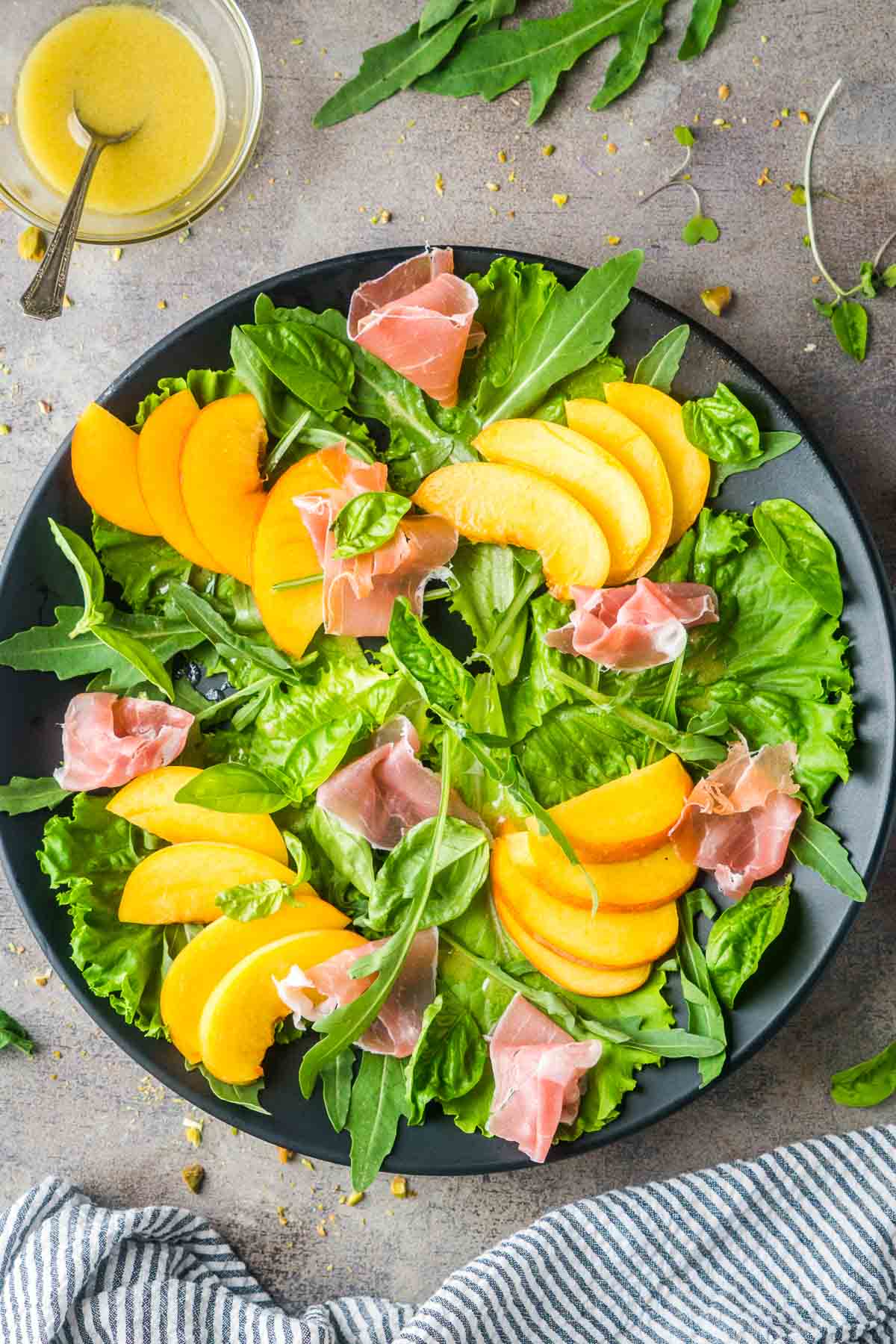sliced peaches, arugula, and prosciutto on black plate next to glass bowl of dressing