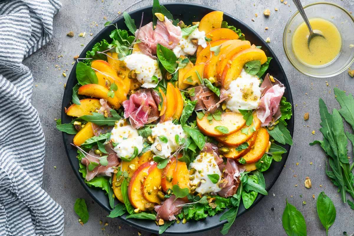 peach burrata salad on black plate next to striped linen and bowl of dressing