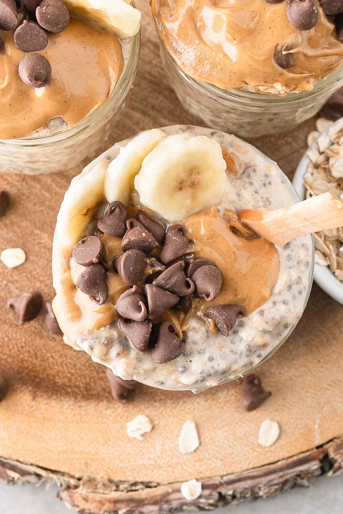 A glass jar filled with peanut butter overnight oats, topped with peanut butter, banana slices, and chocolate chips, sits on a wooden surface next to oats and other jars.