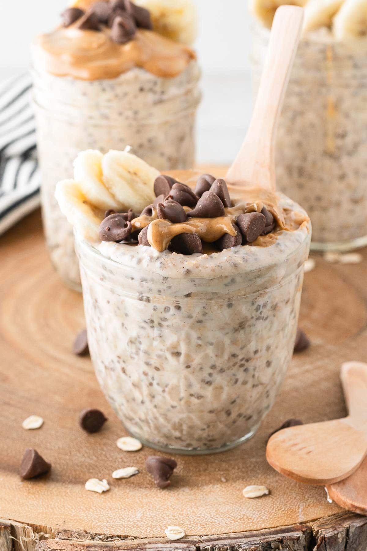 A glass jar filled with peanut butter overnight oats, topped with banana slices, chia seeds, and chocolate chips, with a wooden spoon in front of two jars filled with the same overnight oats.