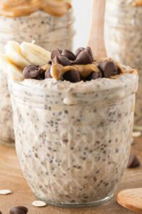 A glass jar filled with peanut butter overnight oats, topped with banana slices, chia seeds, and chocolate chips, with a wooden spoon in front of two jars filled with the same overnight oats.