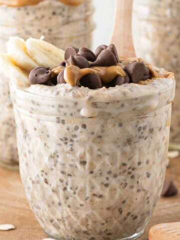 A glass jar filled with peanut butter overnight oats, topped with banana slices, chia seeds, and chocolate chips, with a wooden spoon in front of two jars filled with the same overnight oats.