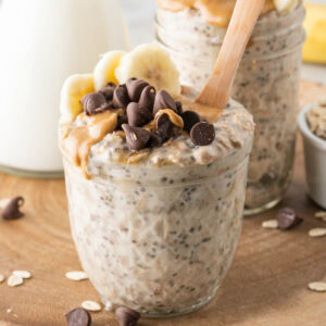 A glass jar filled with peanut butter overnight oats, topped with banana slices, chia seeds, and chocolate chips, with a wooden spoon in front of two jars filled with the same overnight oats.