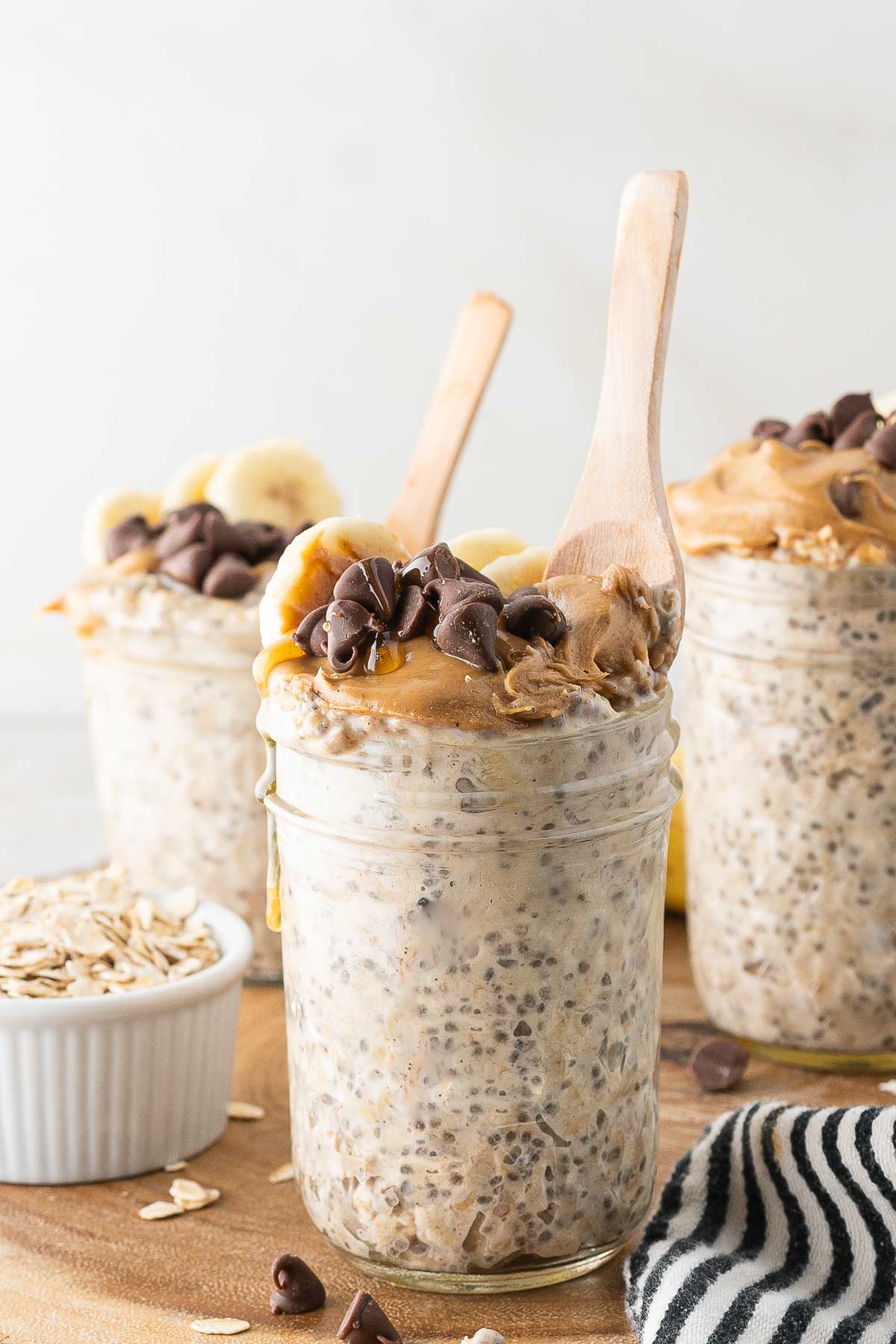 A glass jar filled with peanut butter overnight oats, topped with banana slices, chia seeds, and chocolate chips, with a wooden spoon in front of two jars filled with the same overnight oats.