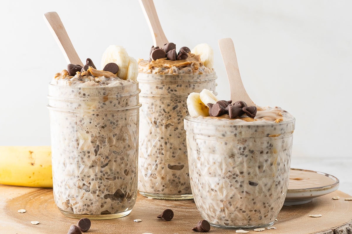 Three jars of peanut butter overnight oats topped with banana slices, chocolate chips, and peanut butter, each with a wooden spoon, are arranged on a wooden surface.