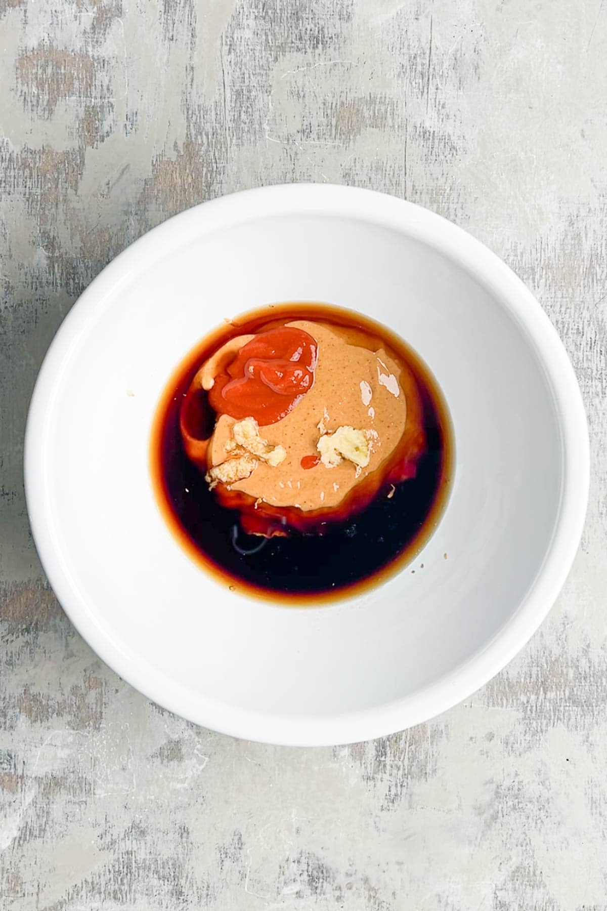 A white bowl containing the base ingredients for peanut sauce&mdash;peanut butter, soy sauce, Sriracha, minced garlic and ginger&mdash;sits on a light textured surface.