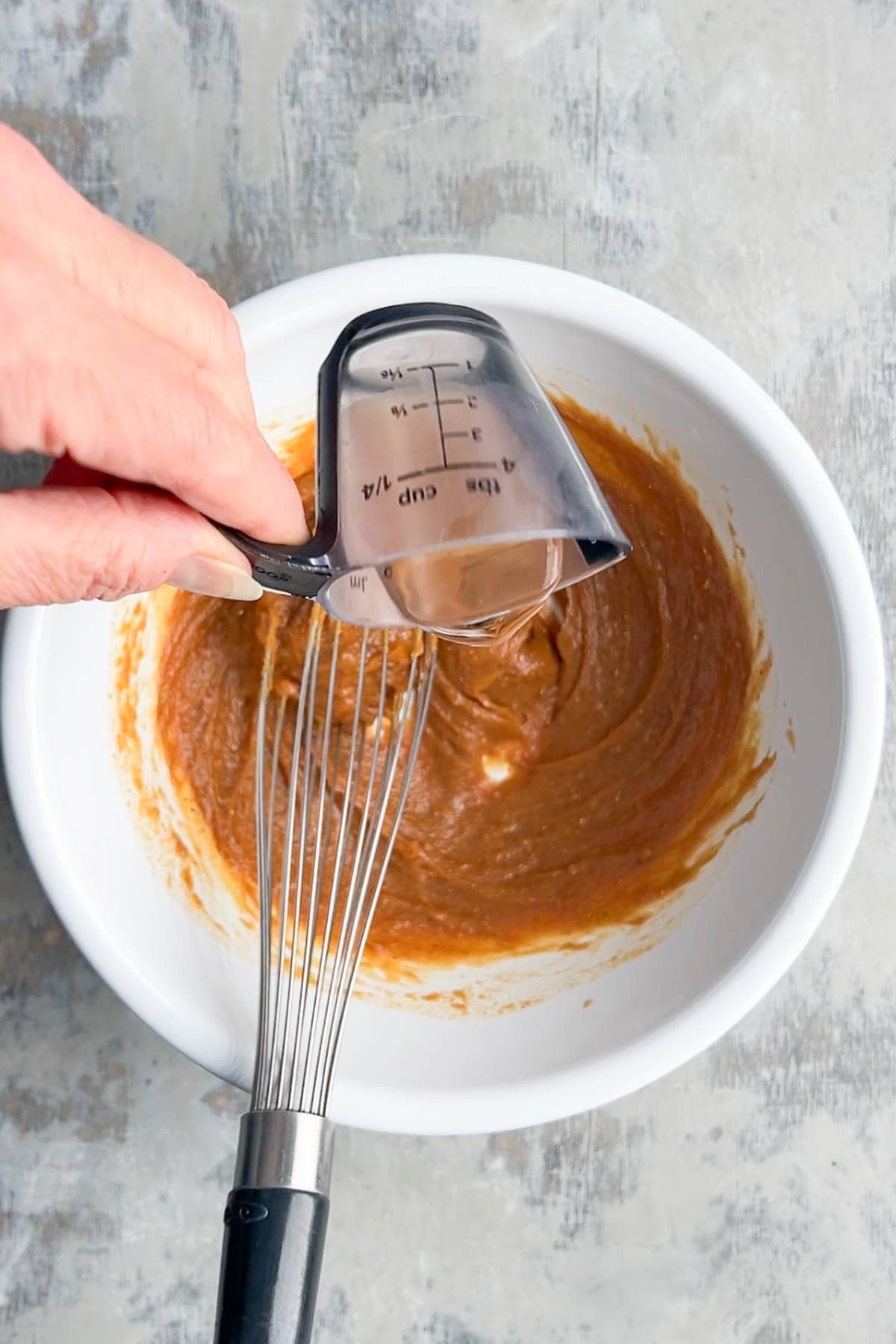 A hand pours liquid from a measuring cup over a white bowl filled with brown peanut sauce mixture and a whisk on a gray surface.