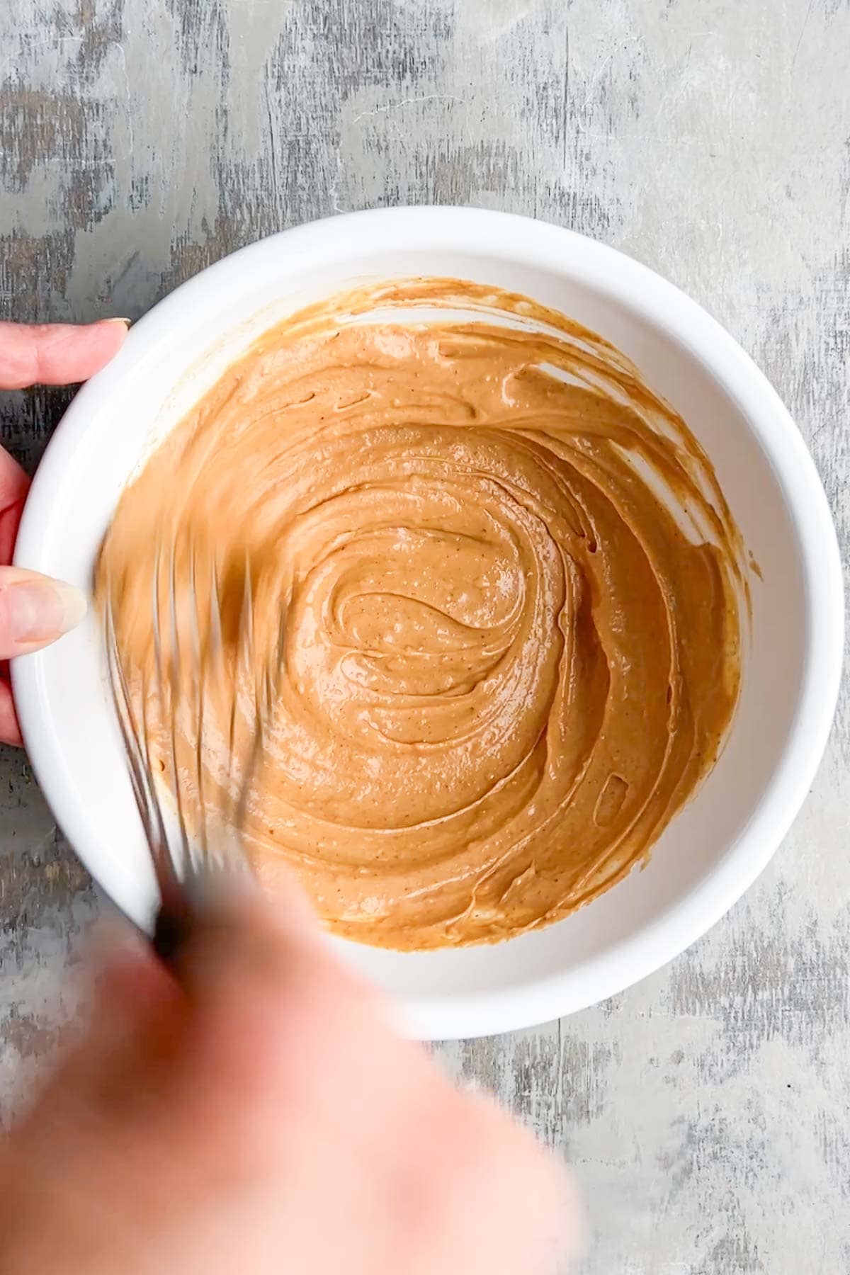 A hand whisking a creamy, light brown peanut sauce in a white bowl on a gray surface.