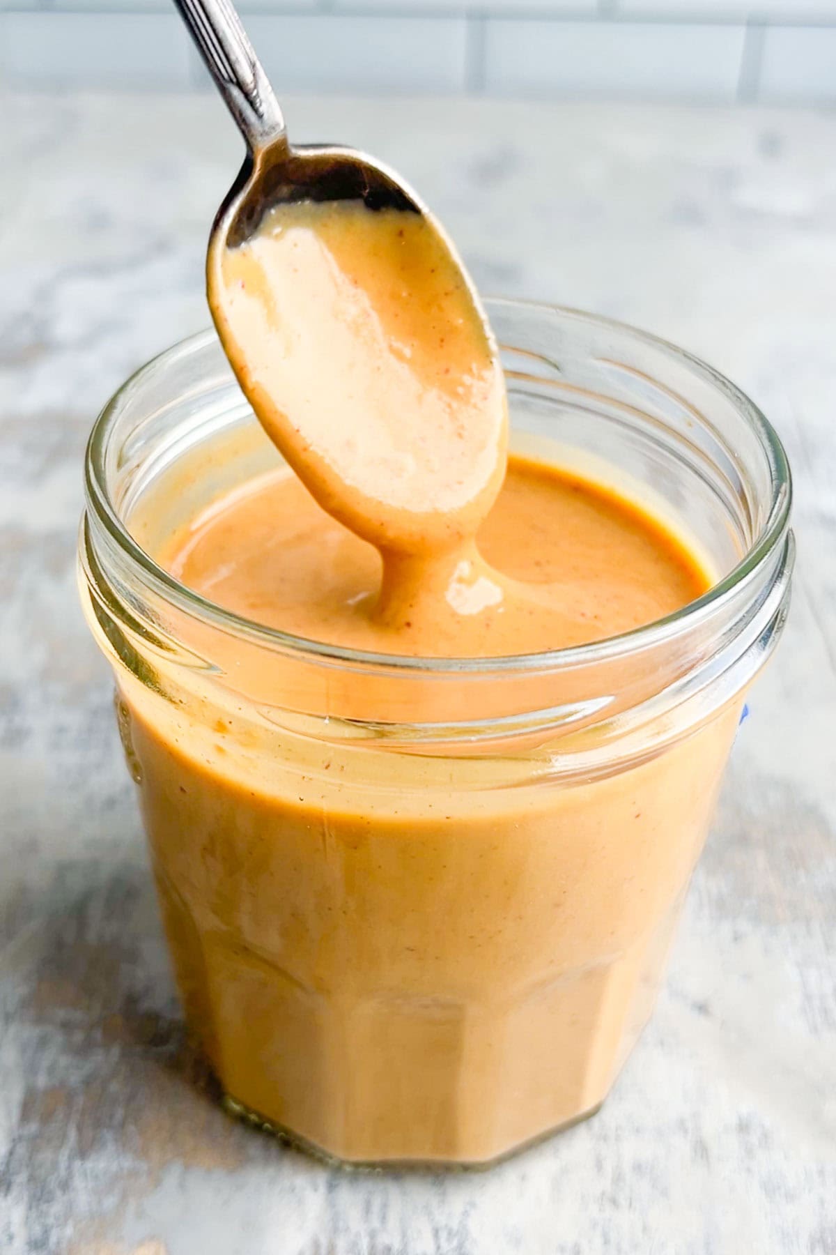A spoon holds up a thick, creamy, orange-brown peanut sauce above a glass jar filled with the same sauce, resting on a light-colored surface.