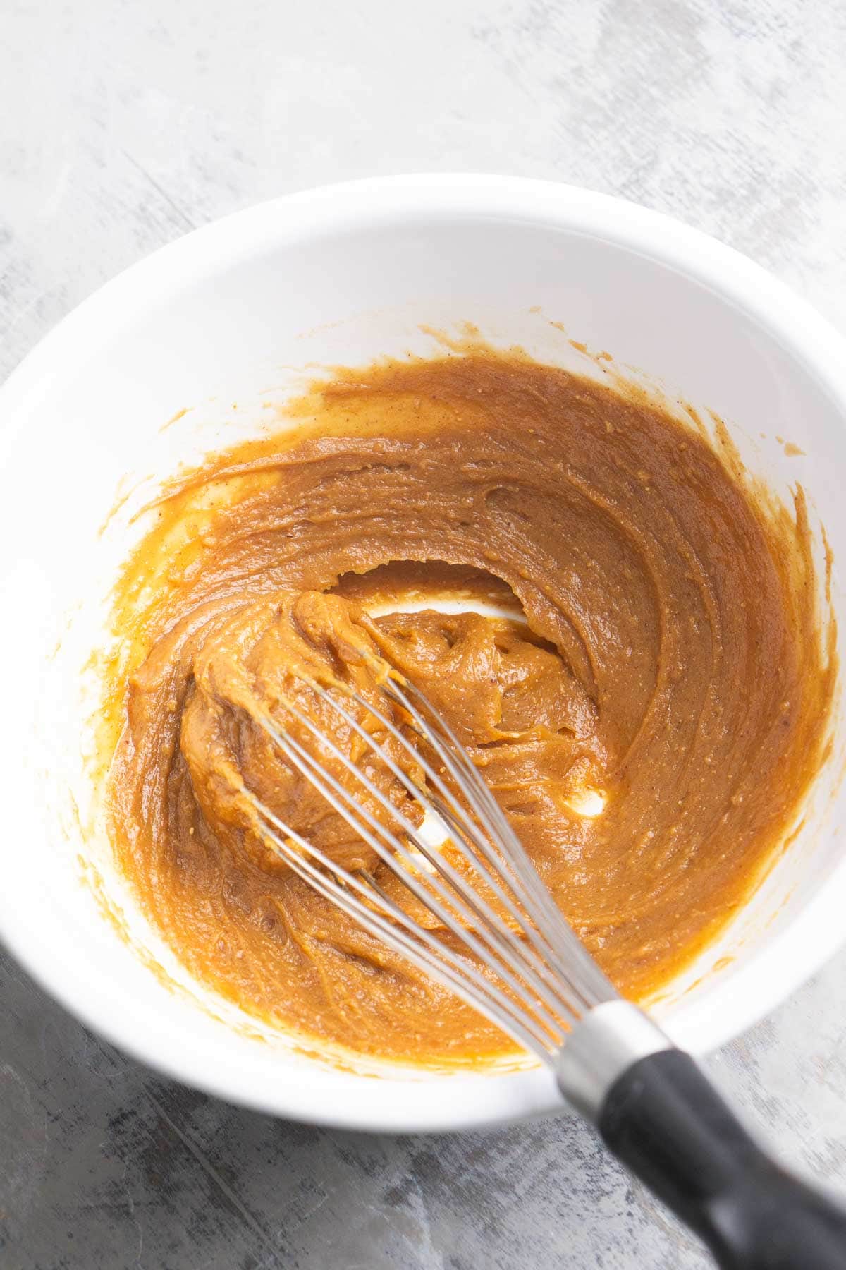 A whisk mixes a thick, brown peanut sauce batter in a white bowl on a light gray surface.