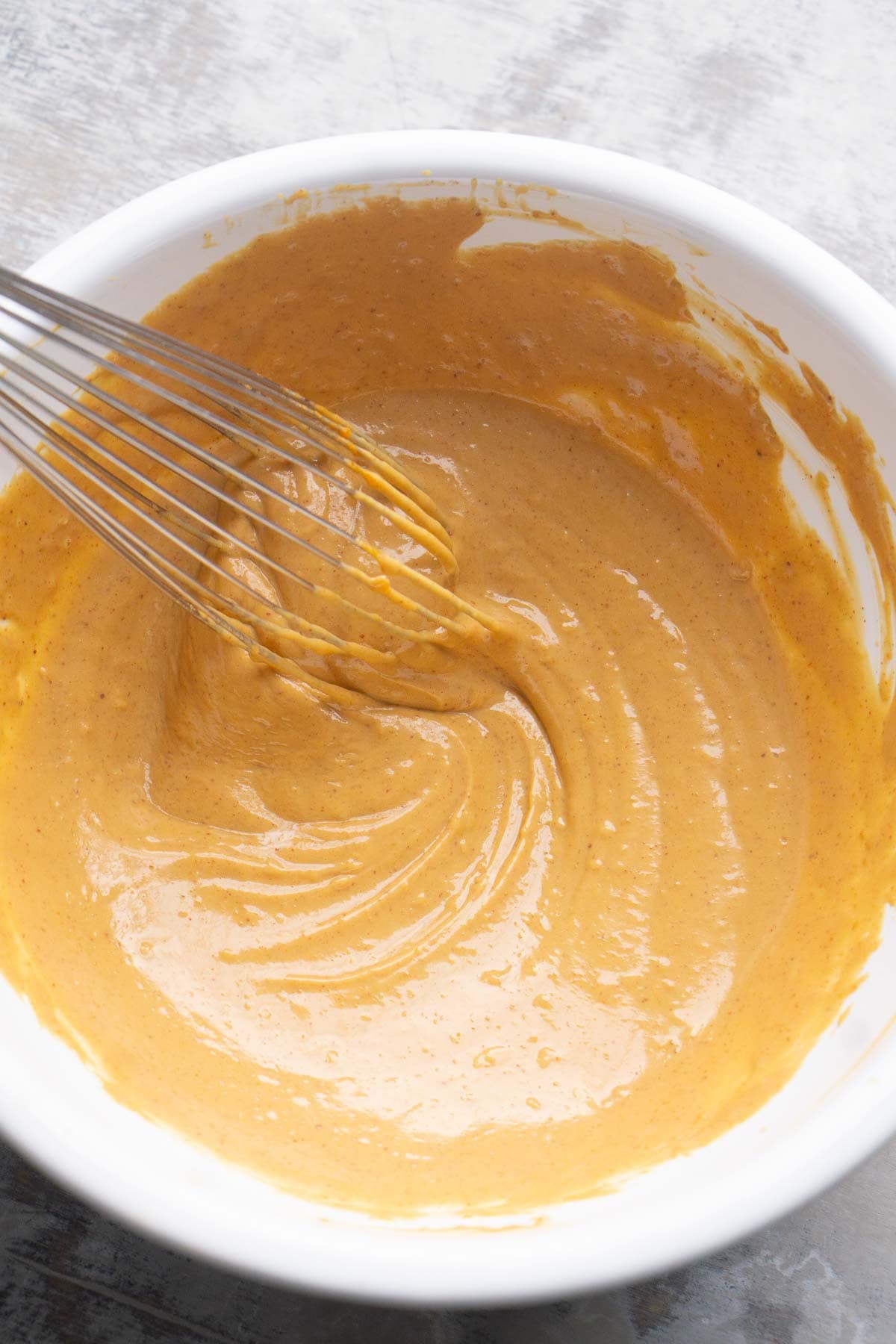 A hand whisking a creamy, light brown peanut sauce in a white bowl on a gray surface.