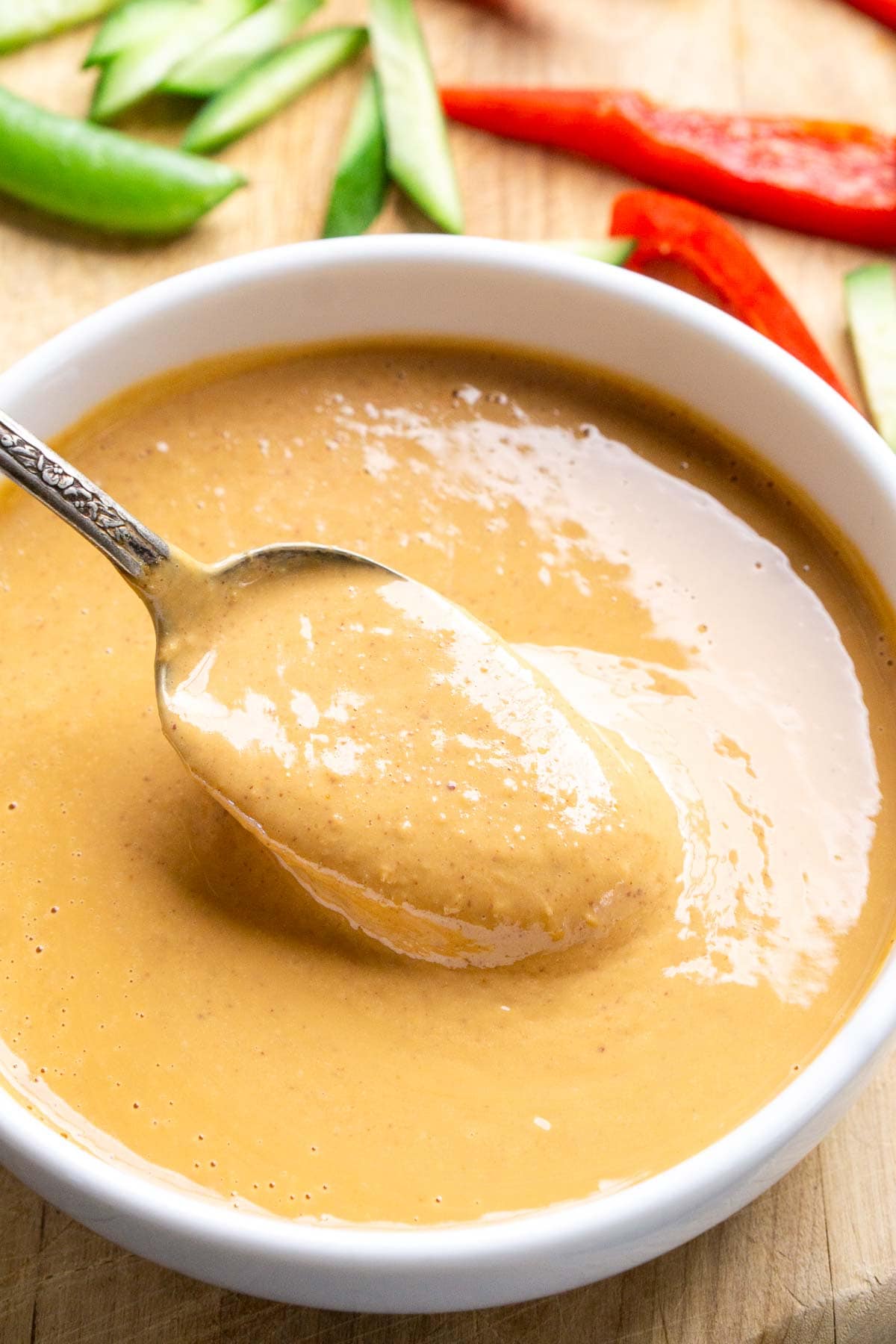 A spoon rests in a bowl of creamy light brown peanut sauce, surrounded by sliced red peppers, cucumber, and snap peas on a wooden surface.
