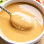A spoon rests in a bowl of creamy light brown peanut sauce in a white bowl.