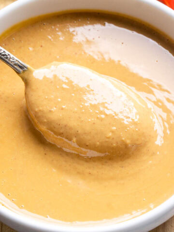A spoon rests in a bowl of creamy light brown peanut sauce in a white bowl.