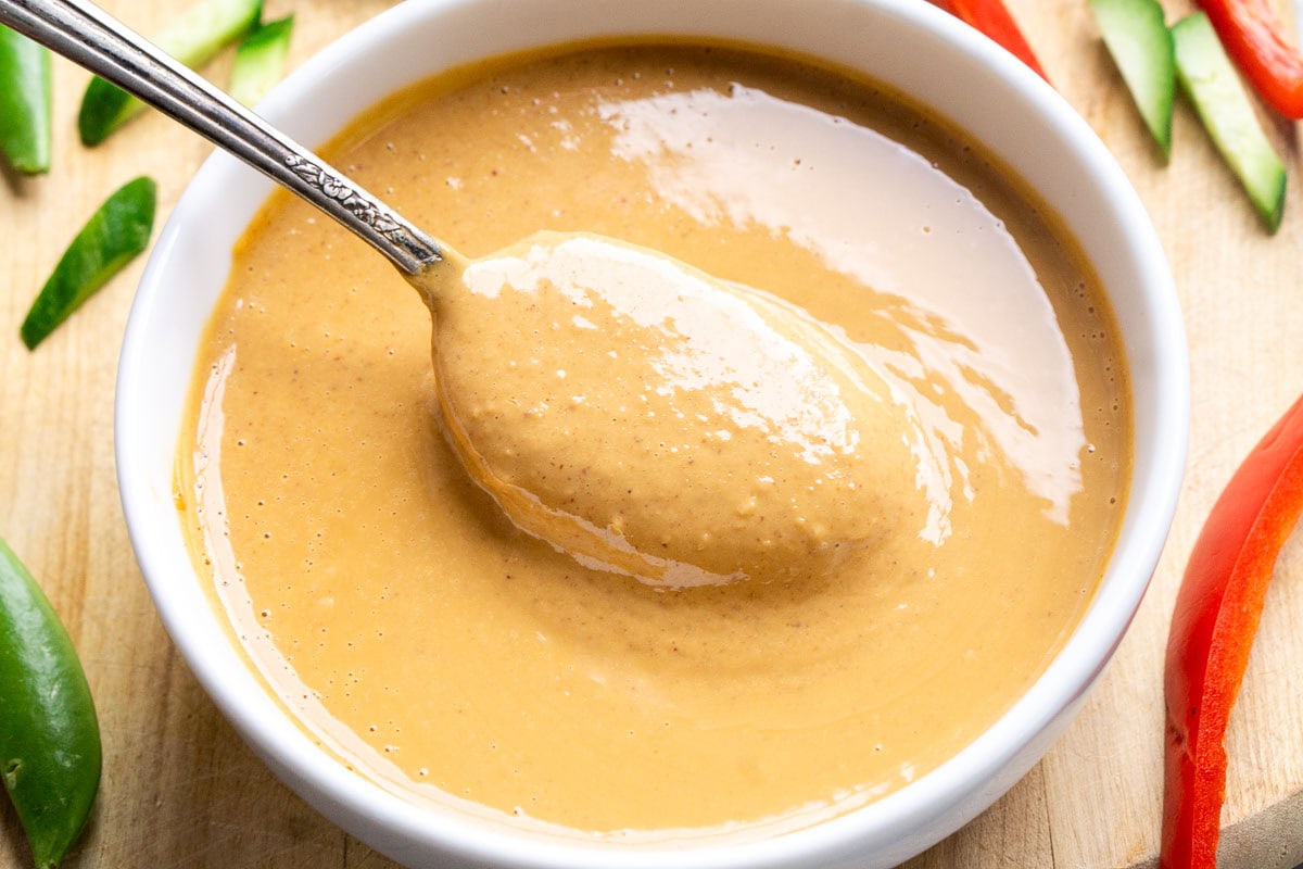 A spoon rests in a bowl of creamy light brown peanut sauce, surrounded by sliced red peppers, cucumber, and snap peas on a wooden surface.