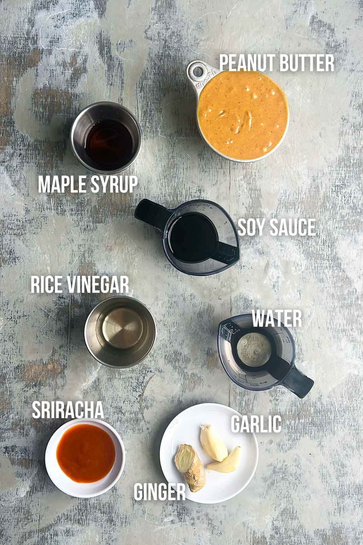 Overhead view of labeled peanut sauce ingredients&mdash;peanut butter, maple syrup, soy sauce, rice vinegar, water, sriracha, garlic, and ginger&mdash;arranged on a gray surface.