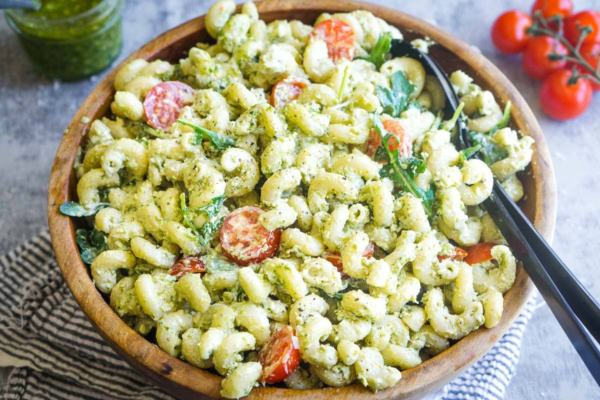A wooden bowl filled with creamy pesto pasta salad mixed with cherry tomatoes, arugula, and salad utensils in the bowl. Nearby, a jar of pesto and vine tomatoes complete the fresh scene.