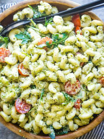 A wooden bowl filled with creamy pesto pasta salad made of cavatappi pasta, cherry tomatoes, and arugula, served with black salad serving utensils.