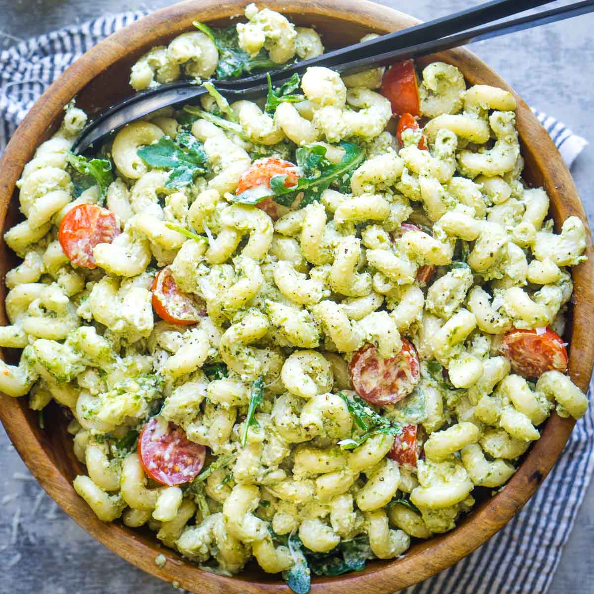 A wooden bowl filled with creamy pesto pasta salad made of cavatappi pasta, cherry tomatoes, and arugula, served with black salad serving utensils.