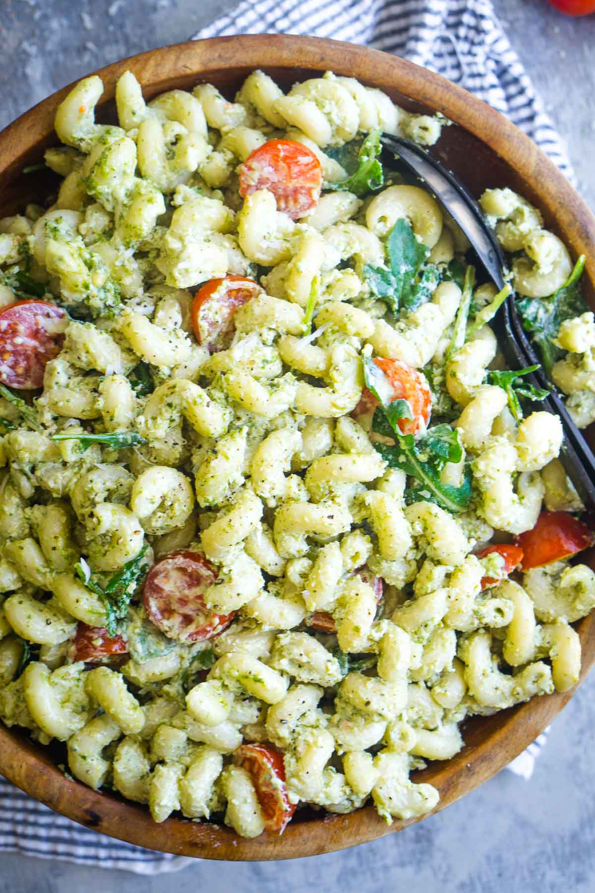 A wooden bowl filled with creamy pesto pasta salad made of cavatappi pasta, cherry tomatoes, and arugula, served with black salad serving utensils.