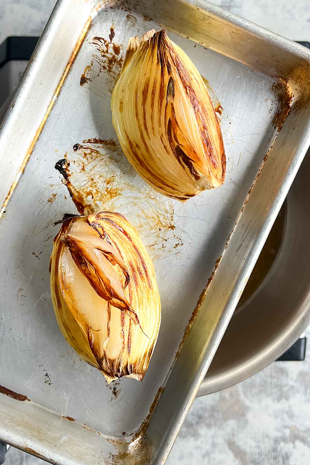 Two roasted onion halves with browned edges on a metal baking sheet.
