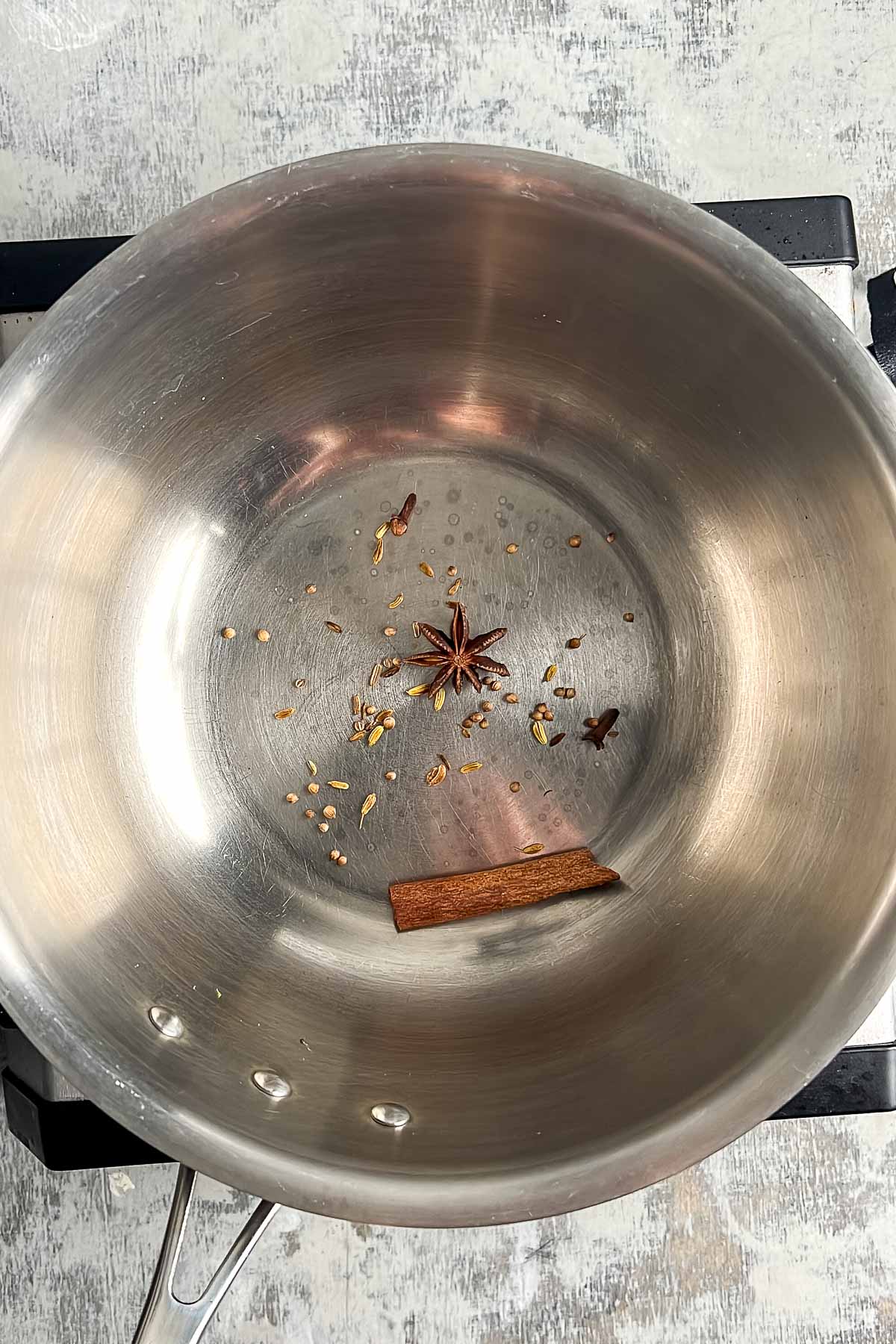 A stainless steel pot with a few whole spices, including a cinnamon stick, star anise, and seeds, sits on a stovetop.