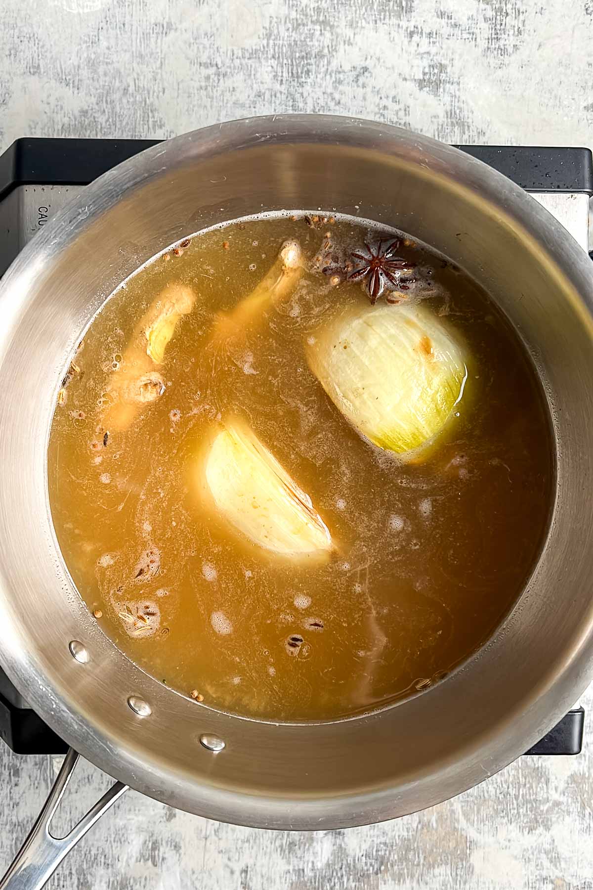 A pot of broth simmers on a stovetop, containing onion halves, ginger pieces, star anise, and other visible ingredients.
