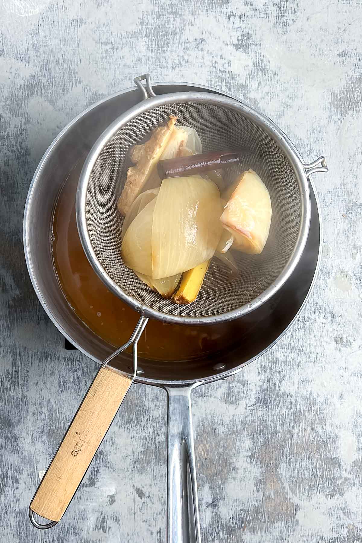 A metal strainer holding cooked onion, ginger, and cinnamon stick sits over a pot with rich brown pho noodle soup broth on a gray surface.